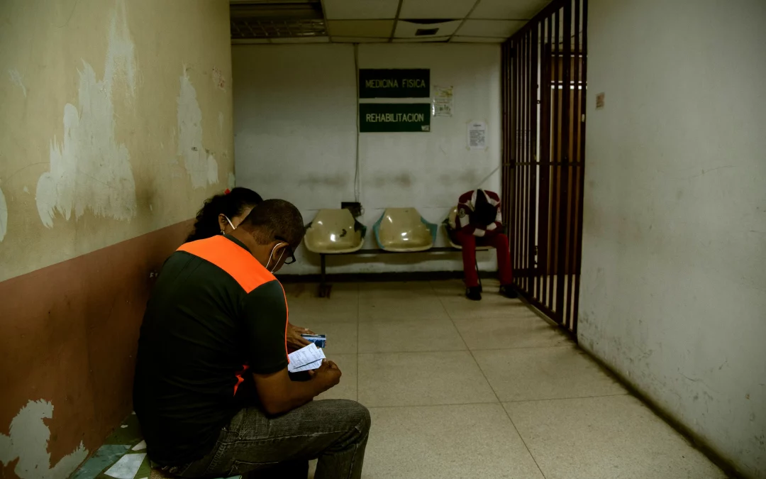 Dos personas sentadas en la sala de espera de un hospital en Venezuela con poca iluminación y de fondo una persona con la cabeza sentada entre las rodillas.