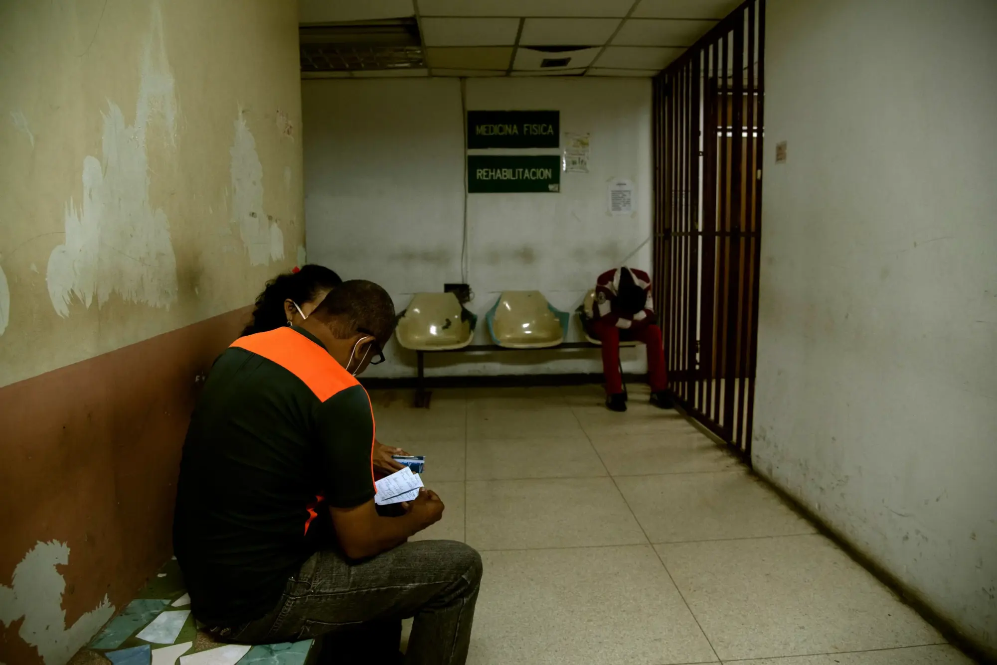 Dos personas sentadas en la sala de espera de un hospital en Venezuela con poca iluminación y de fondo una persona con la cabeza sentada entre las rodillas.