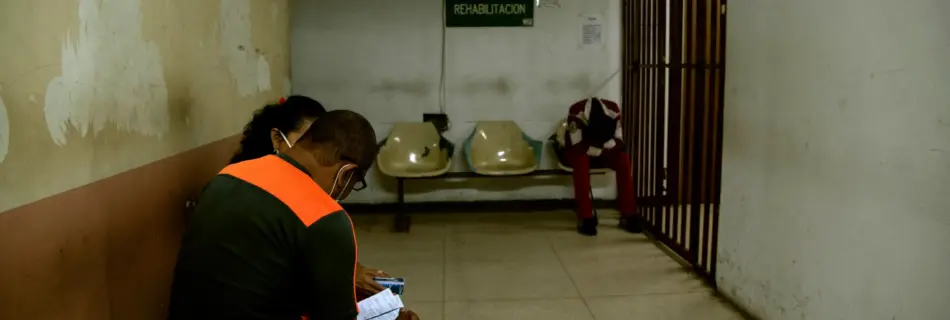 Dos personas sentadas en la sala de espera de un hospital en Venezuela con poca iluminación y de fondo una persona con la cabeza sentada entre las rodillas.